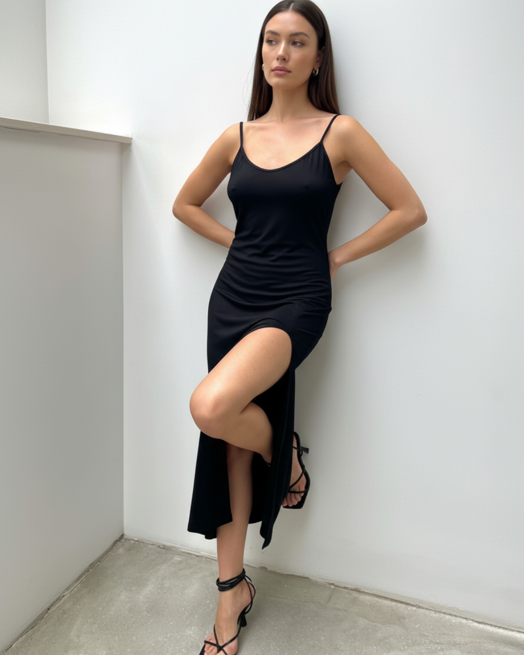 Czarna długa sukienka na ramiączkach Stella z wysokim rozcięciem na nogę. Modelka w czarnej sukience maxi oparta o ścianę, stylizacja wieczorowa z sandałkami.